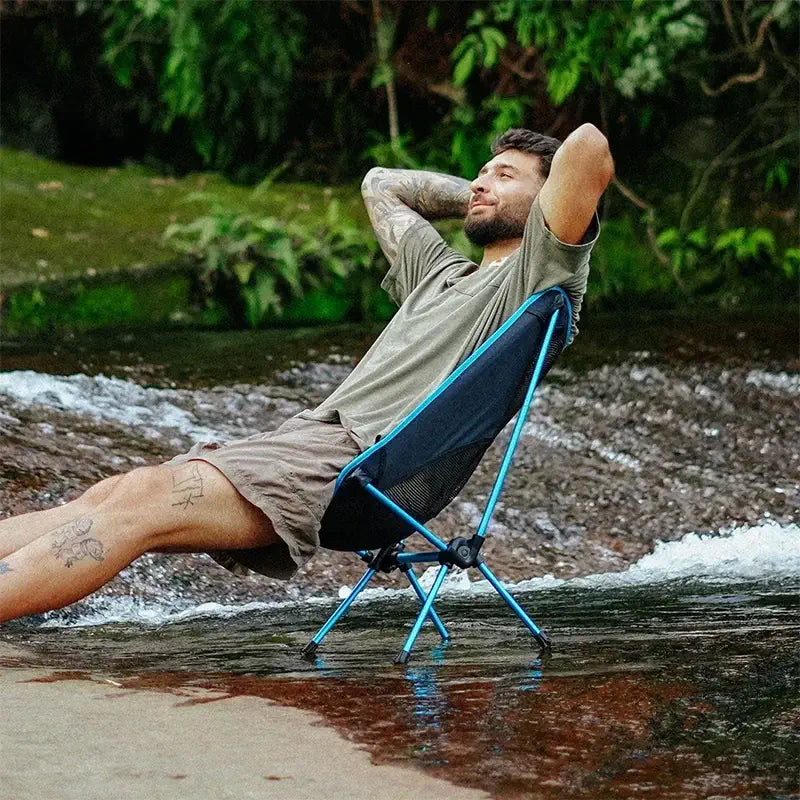 Homem sentado confortavelmente na cadeira portátil azul relaxando na beira do rio