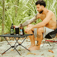 Homem utilizando a mesa portátil na areia, demonstrando a estabilidade e conveniência para uso em praia e ambientes externos.