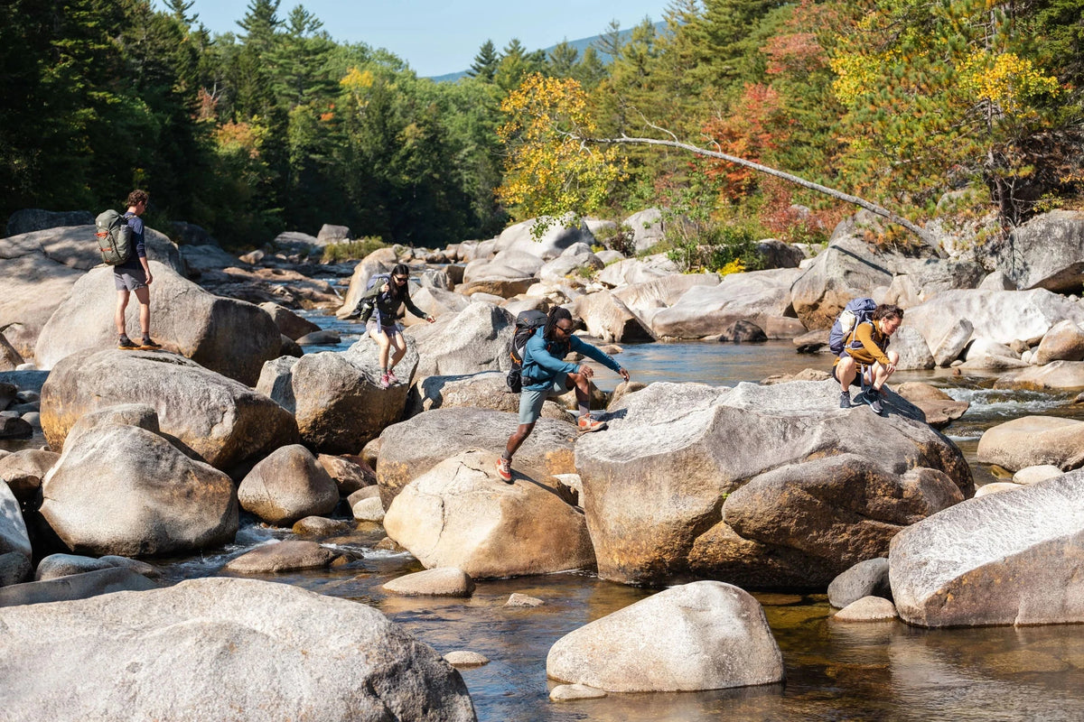 Grupo de aventureiros atravessando pedras em rio de montanha — trilhas e mochilas outdoor da Cultura Puff