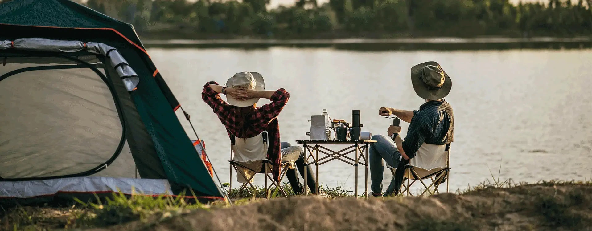 Dois aventureiros relaxando em cadeiras dobráveis à beira de um lago, com barraca montada ao lado e pôr do sol ao fundo — cena de camping e tranquilidade.